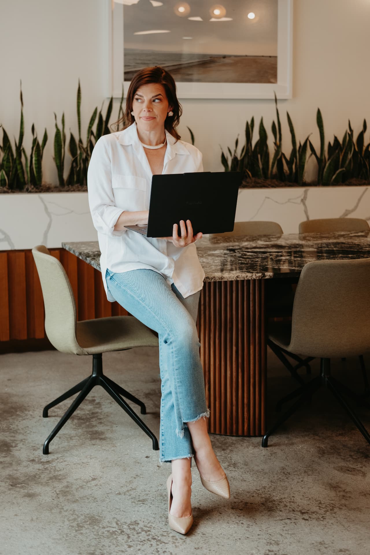 Kellena in a white shirt and jeans, seated on a marble table holding a laptop, looking to the side
