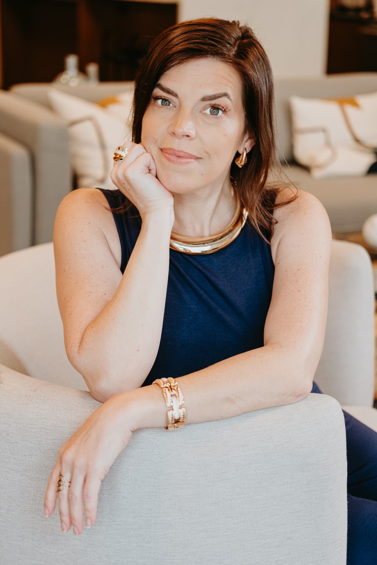 Kellena seated on a light sofa, chin resting on her hand, in a navy dress with gold jewellery